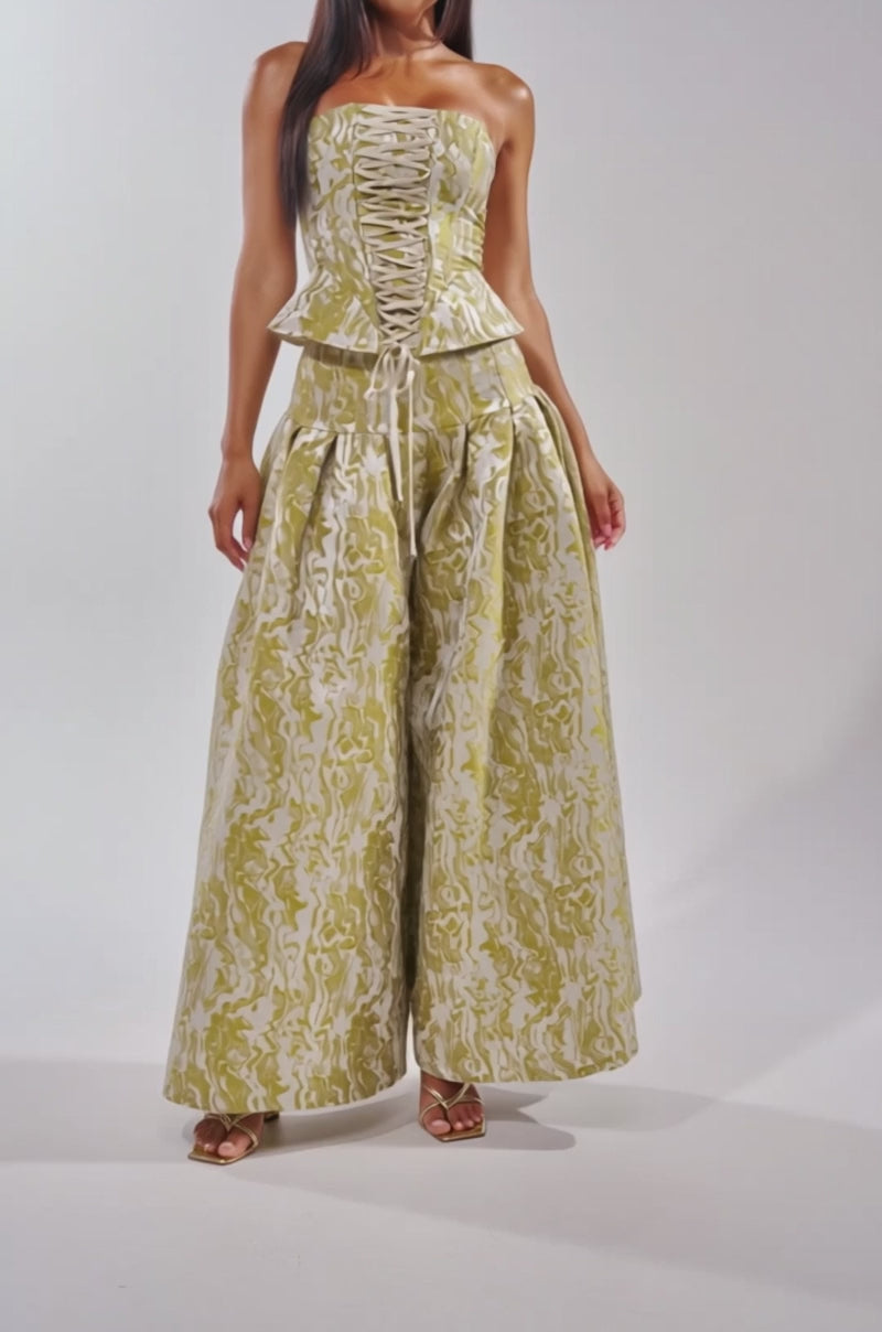 A video of a woman wearing sandals and the GET IT FLORAL TROUSER—a green brocade pant with a textured pattern—paired with a strapless, lace-up peplum top.