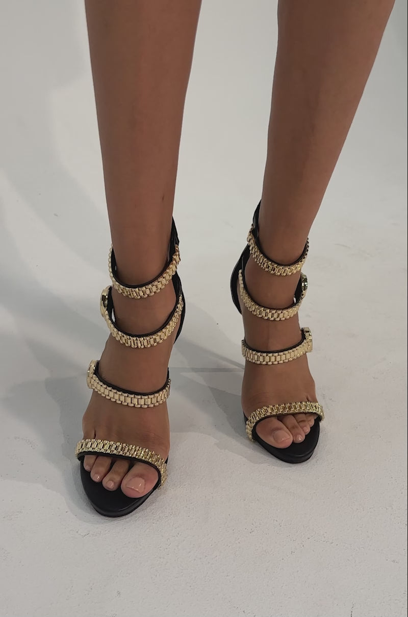 A video of a person wearing the AZALEA WANG BEZEL WATCH STRAP NOVELTY STILETTO SANDAL—black heels with gold-tone watch-link straps and coin embellishments.