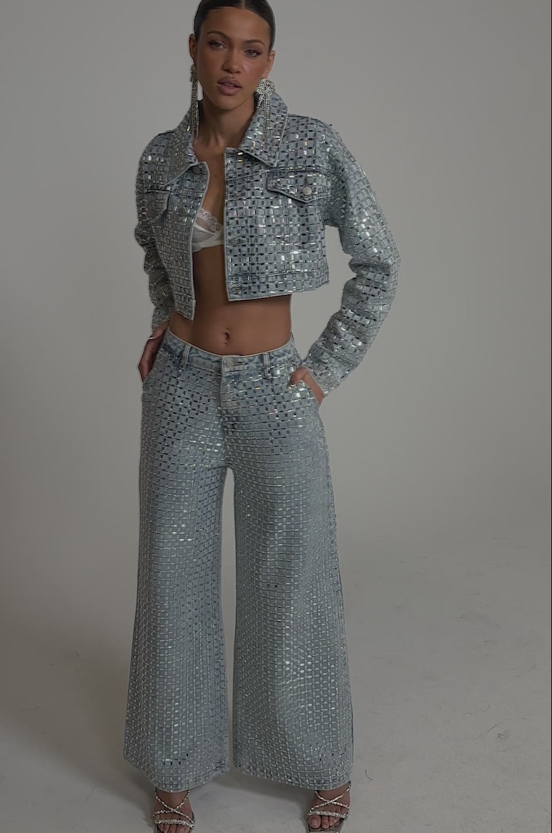 A video of a woman wearing PICK N CHOOSE RHINESTONE WIDE LEG DENIM pants with a shiny, light blue sequin cropped jacket, white lace bralette, and heeled sandals, posing against a plain background.