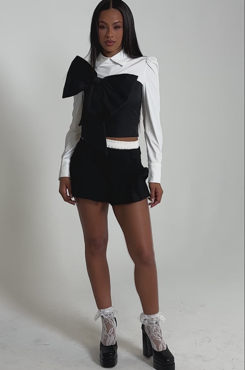 A video of a woman wearing the JACQUELINE POPLIN BLOUSE with a white collar, black shorts, white lace socks, and black high-heeled platform shoes.