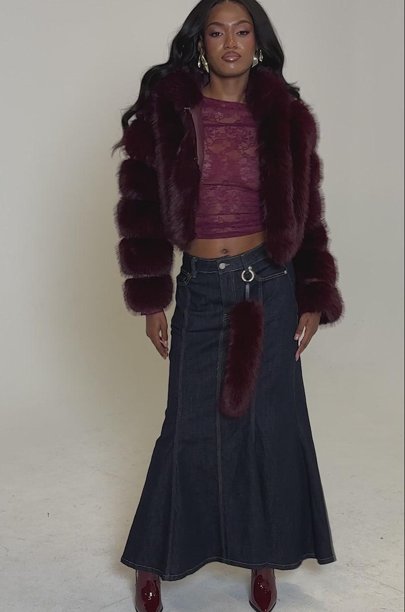 A video of a woman posing against a plain background in the JOLENE FAUX FUR COAT IN BURGUNDY, styled over a sheer lace top, a long dark denim skirt, matching burgundy heeled boots, and a furry accessory hanging from her waistband.