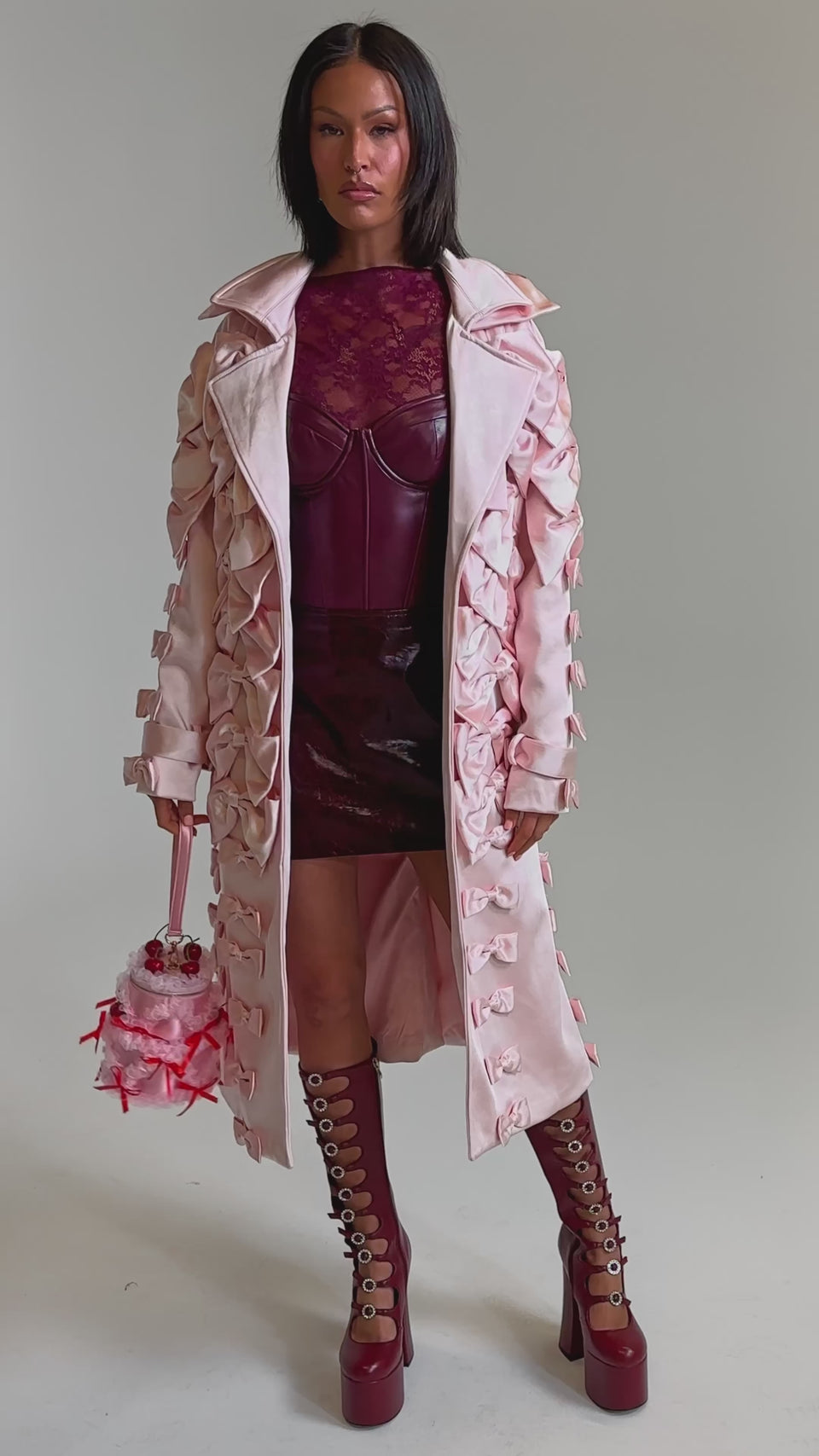 A video of a woman in the WRAP ME WITH A SATIN BOW TRENCH, styled with a burgundy lace top, matching mini skirt, tall burgundy platform boots with grommets, and a pink handbag featuring red ribbons and bows.