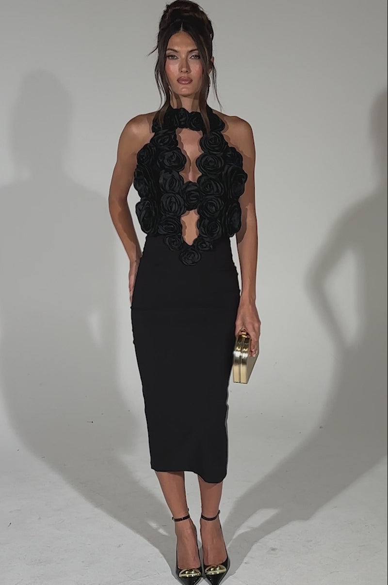 A video of a woman posing in the ALWAYS ON THE GUEST LIST MIDI DRESS, a fitted black midi with a halter neckline and bold 3D black fabric roses, holding a gold-accented clutch against a plain light background. 