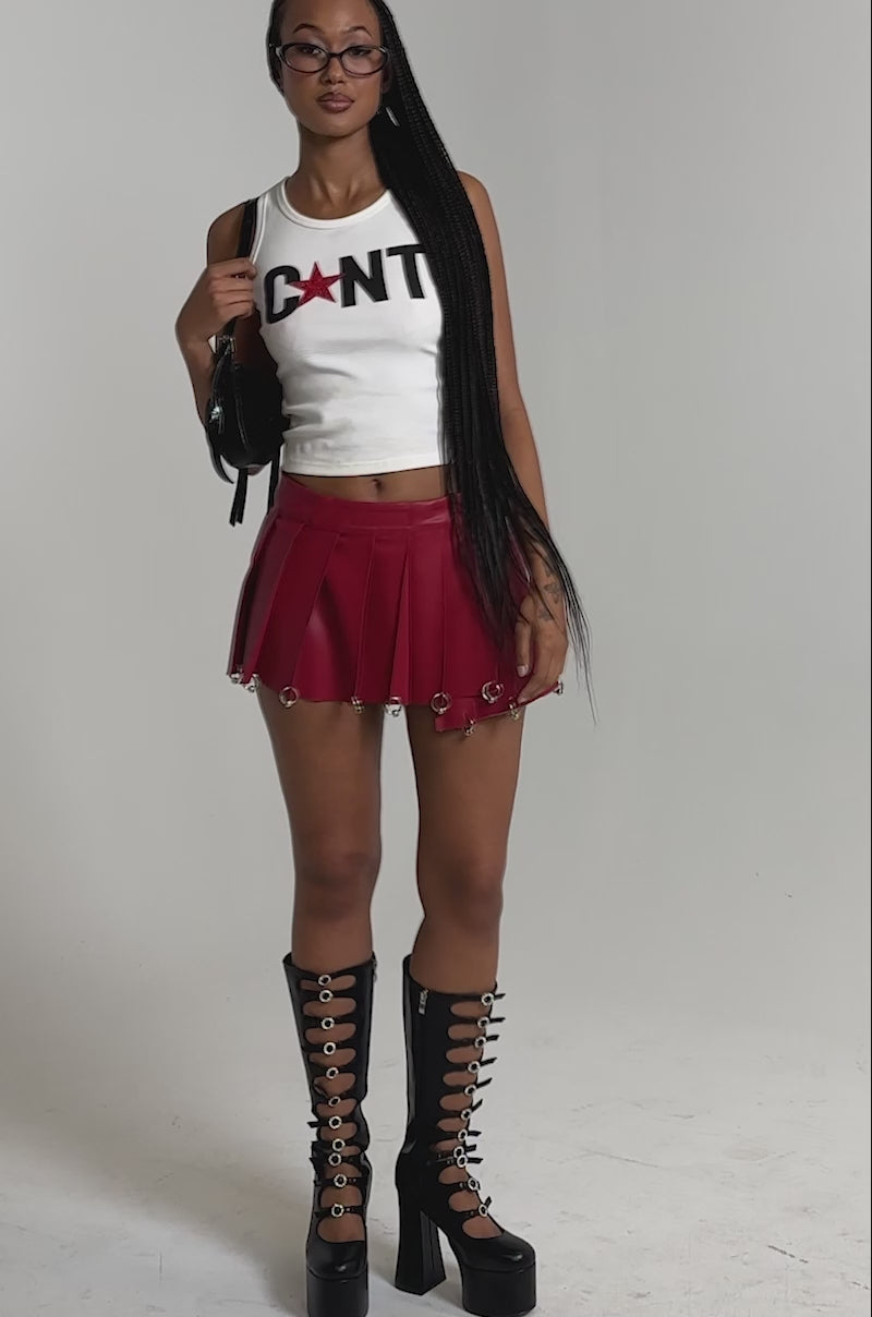 A video of a woman wearing glasses, a CNTY LACE UP TANK, a short red pleated skirt, black knee-high platform boots with buckles, and holding a black handbag with a bow.