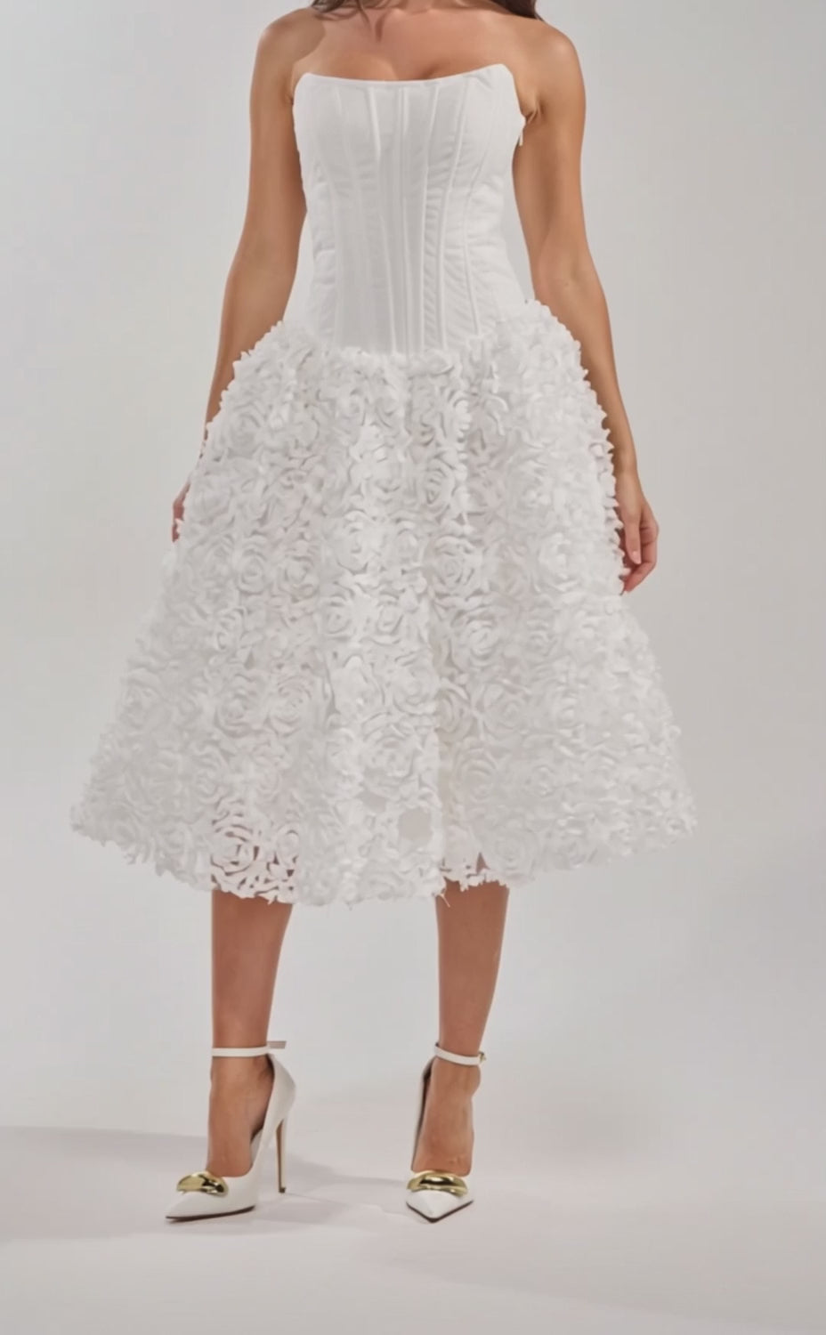 A video of a woman wearing the FIND ME LATER FLORAL CORSET MIDI DRESS IN WHITE, featuring a strapless fitted bodice and voluminous skirt, paired with gold pointed-toe heels.