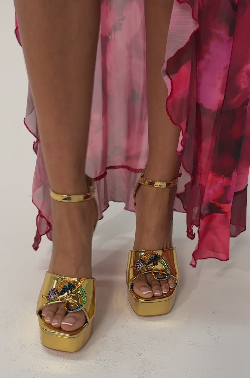 Video of woman modeling the AZALEA WANG PASQUALE GOLD IGUANA EMBELLISHED SANDAL—metallic gold platform heels adorned with colorful lizard decorations—while standing on a light surface.