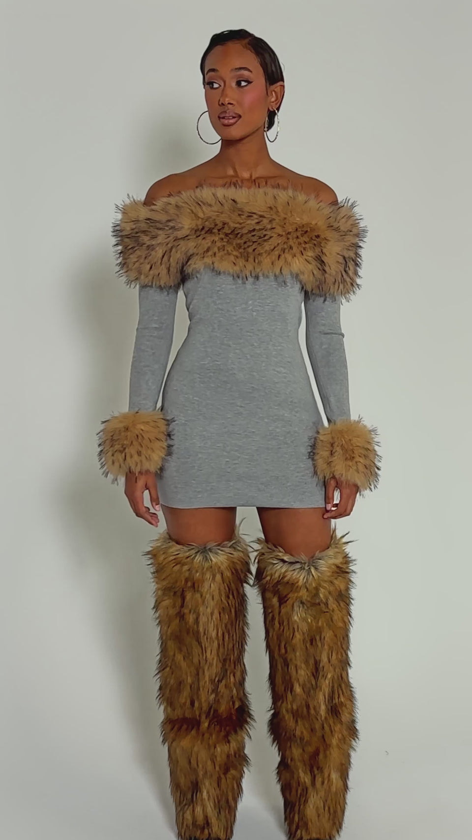 Video of woman modeling the COMFY COZY FUR TRIM MINI DRESS IN GREY, featuring a fitted ribbed knit, off-shoulder cut, and fur trim on the neckline and cuffs, paired with matching boots against a light background.