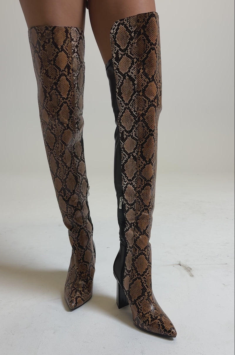 A video of a person standing on a neutral background wearing brown faux leather shorts and AKIRA GOLD NOAMI MULTI SNAKE PRINT CHUNKY HEEL BOOTS.