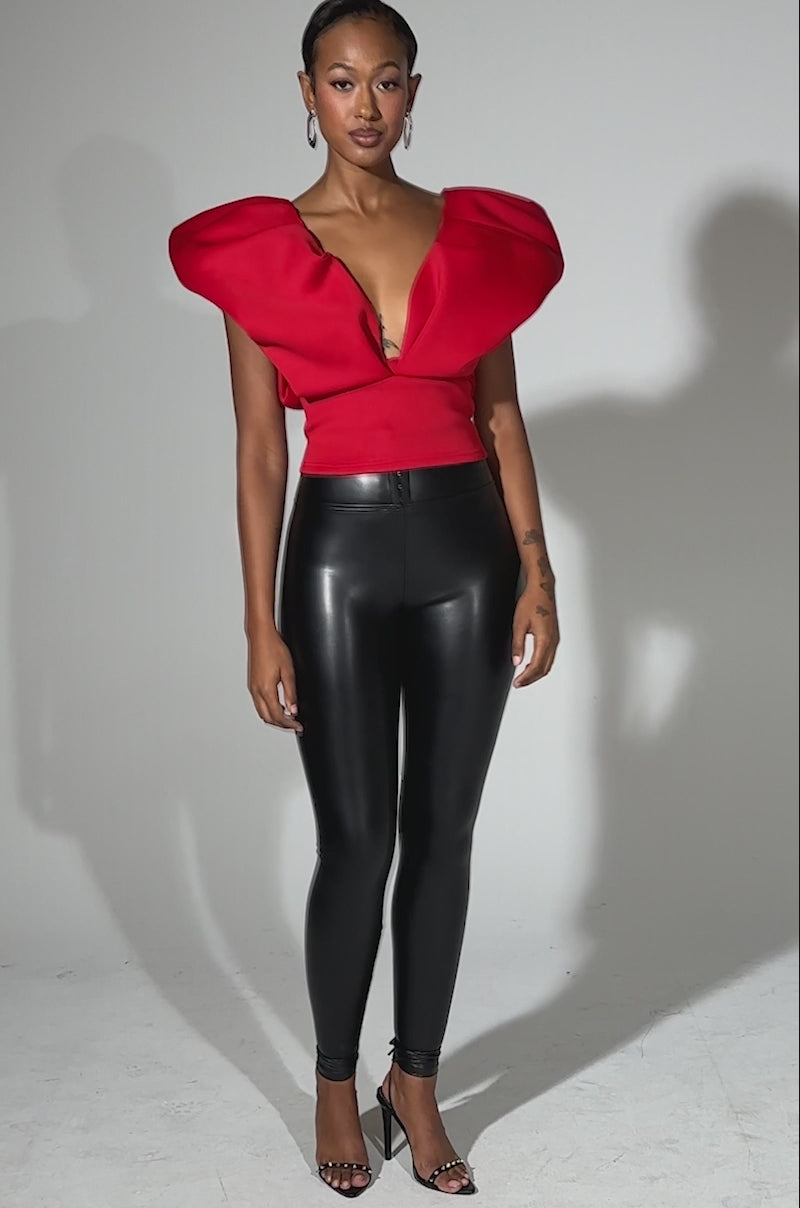 A video of a woman posing confidently in the FEMME ENERGY SCUBA BLOUSE IN RED, featuring sculpted shoulders, paired with black high-waisted leather leggings and black open-toe heels against a plain light background.