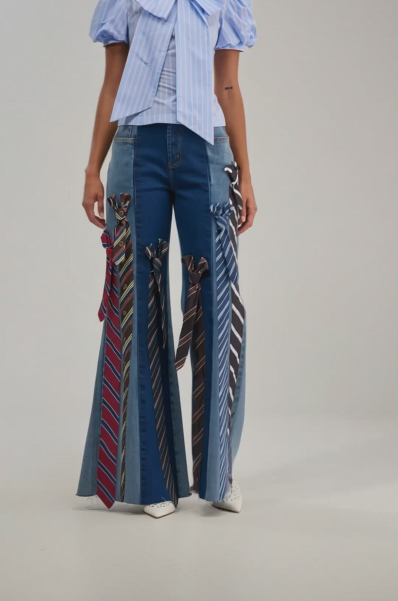 A video of a woman wearing the TIED TO YOU RELAXED DENIM—wide-leg jeans with neckties on the legs—paired with a blue striped blouse featuring puffed sleeves and a large neck bow.