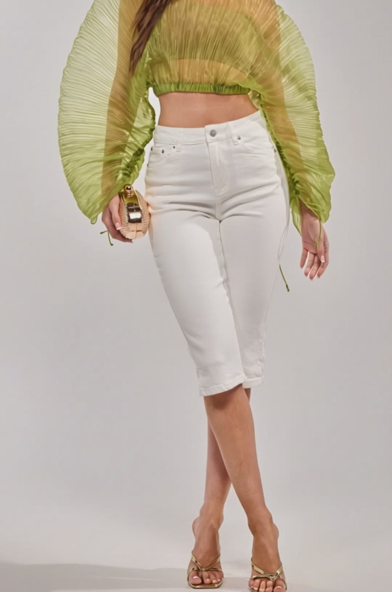 A woman is wearing the CICI DENIM CAPRI, a light green sheer pleated-sleeve top, gold heeled sandals, and holding a small textured clutch stands against a plain light background.