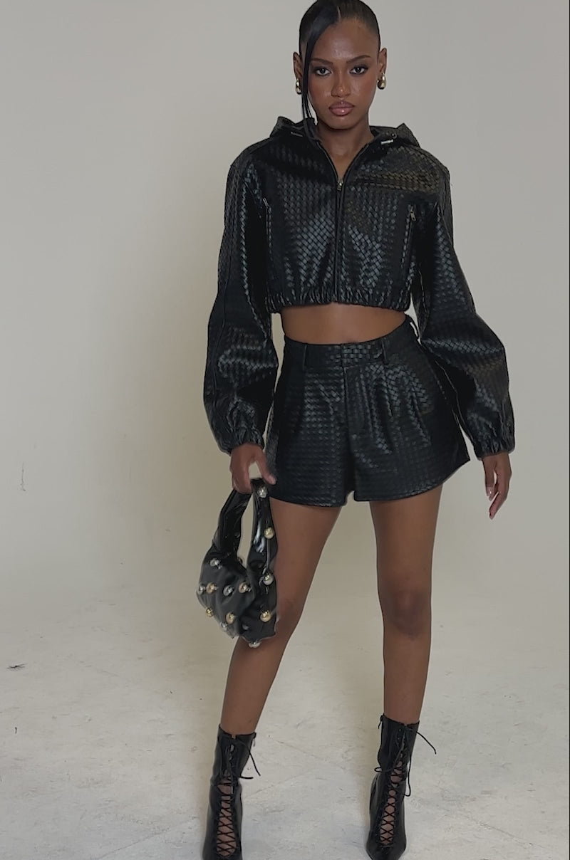 Video of woman modeling the CHANGE OF PERSPECTIVE FAUX LEATHER SHORT with a matching black cropped jacket, lace-up ankle boots, and a black handbag. The setting is minimal, with a plain, light-colored background.