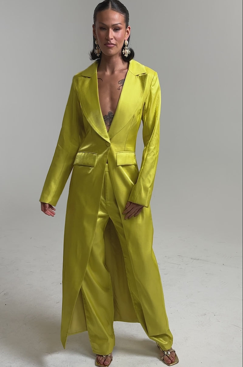A video of a person in the CLEO TRENCH IN CHARTREUSE with matching wide-leg pants, styled with strappy heels, statement earrings, and layered necklaces against a neutral background.