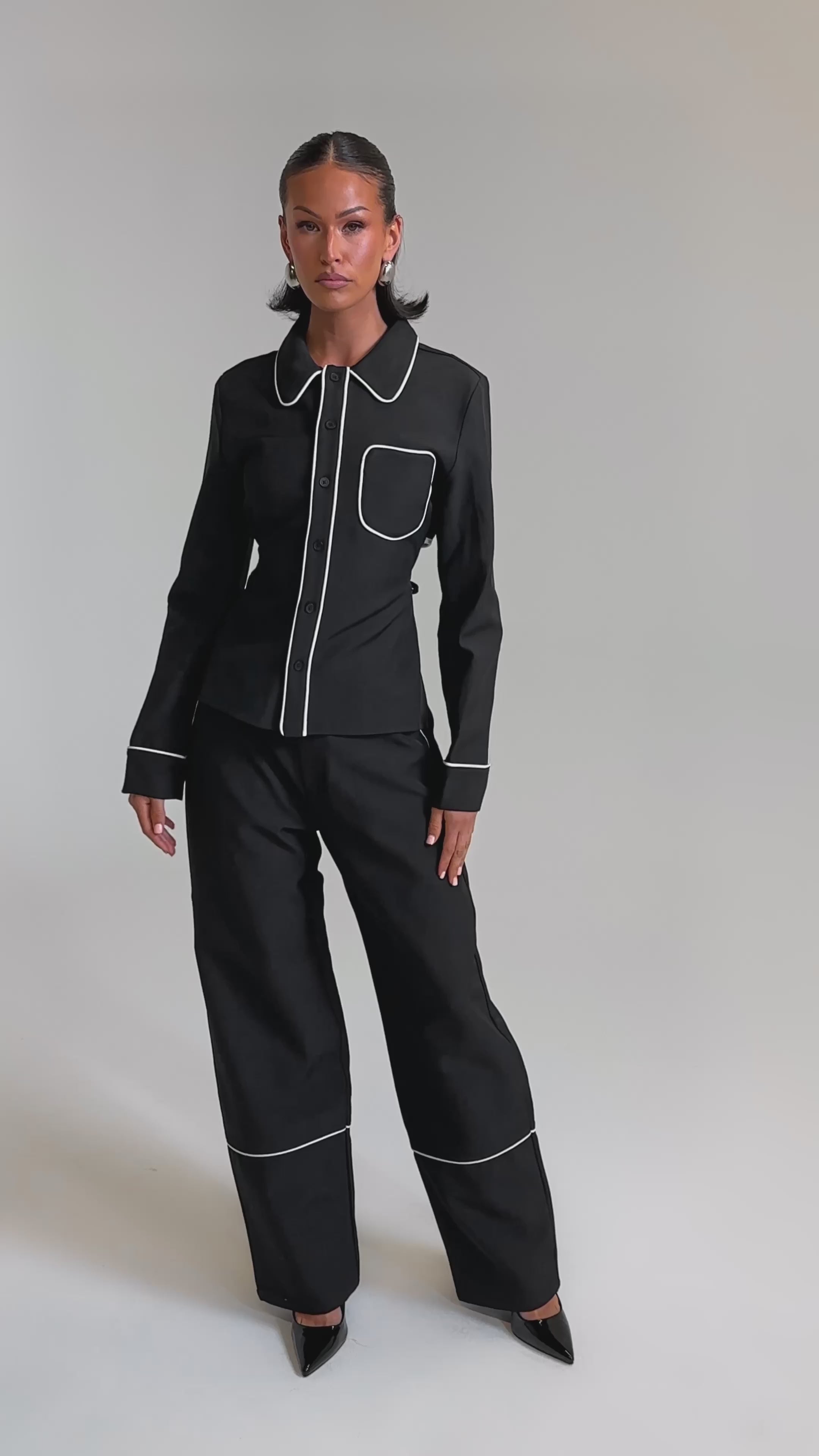 A video of a woman in the PARTY AT MY PLACE TROUSER, a tailored black high-rise wide-leg pant with white piping, paired with a matching collared jacket featuring a chest pocket. She completes the look with black pointed-toe heels and slicked-back hair.