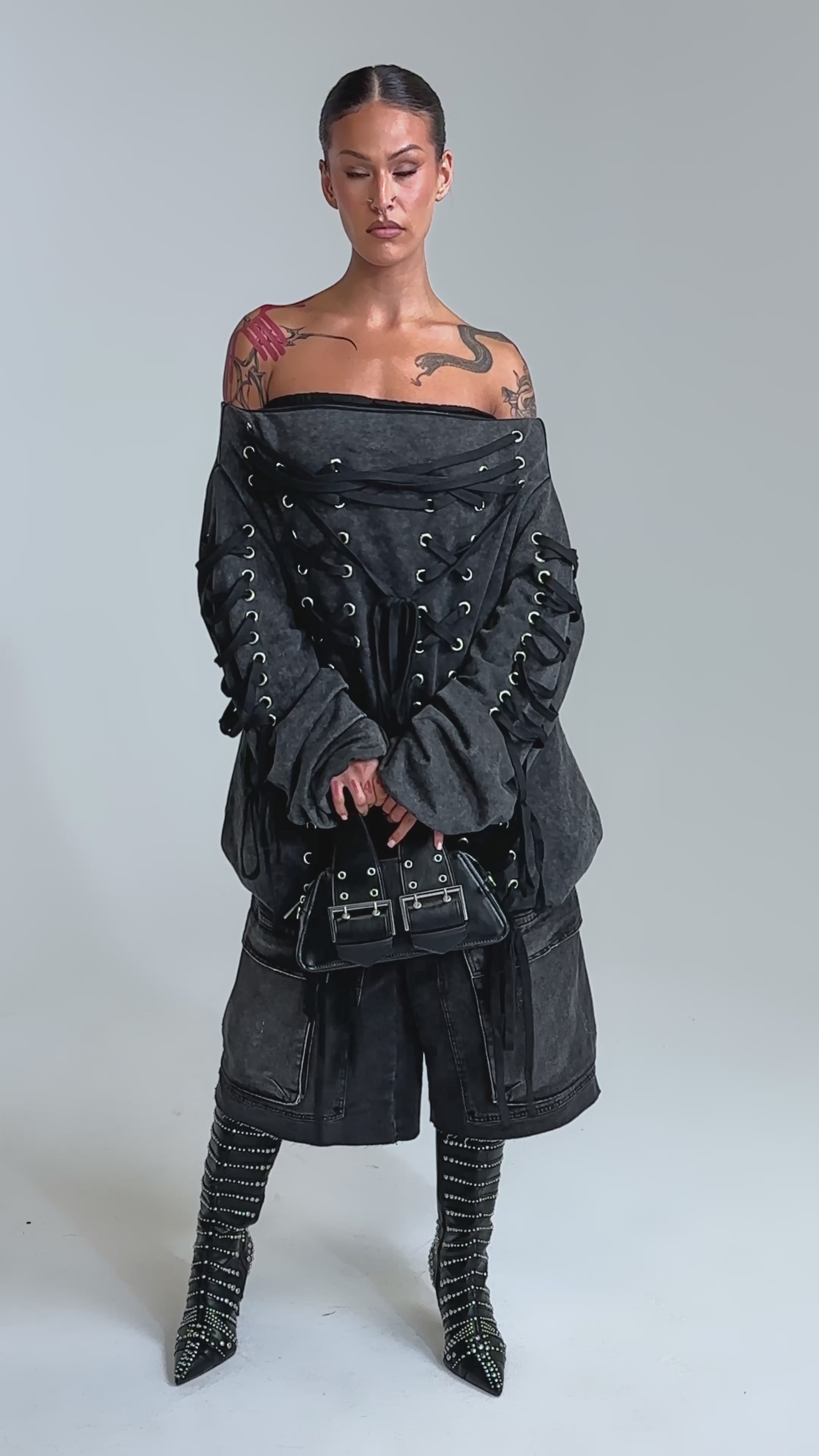Video of woman modeling the LACED UP FOR GOOD MINI DRESS, a charcoal gray, oversized off-the-shoulder piece with lace-up detailing and wide sleeves, paired with baggy black pants and a black handbag.