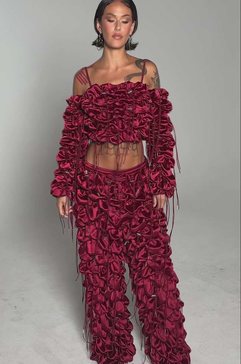 A video of a woman standing in the FEELING FUNKY TIE BLOUSE, a bold maroon off-the-shoulder top with textured ruffled layers and thin ties, paired with matching wide-legged pants against a plain gray background.