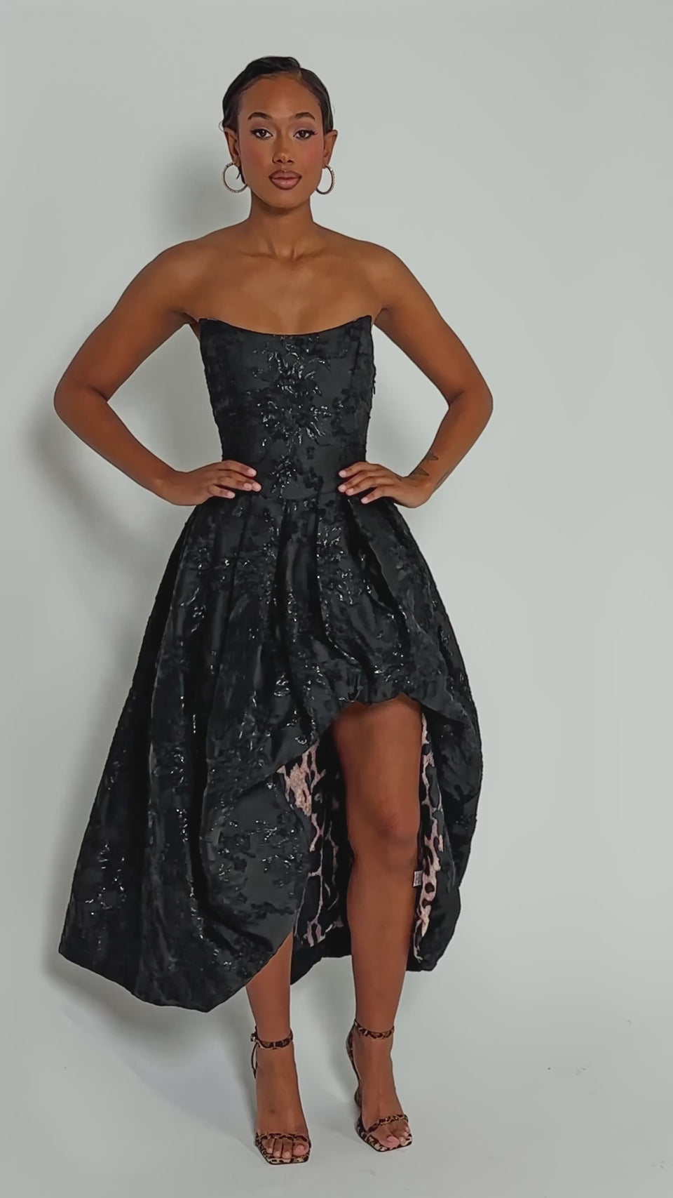 Video of woman modeling the WILD THANG PRINTED HI LOW DRESS, featuring a strapless jacquard floral pattern with leopard print lining. She completes the look with hoop earrings, strappy heels, and styled-back hair for an elegant finish.
