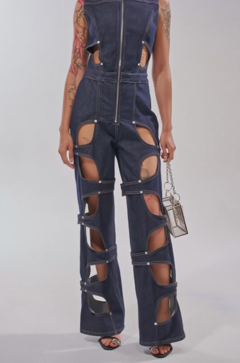 Video of woman modeling the BRING THE ENERGY CUT OUT DENIM JUMPSUIT, paired with black heeled sandals and a metallic handbag.