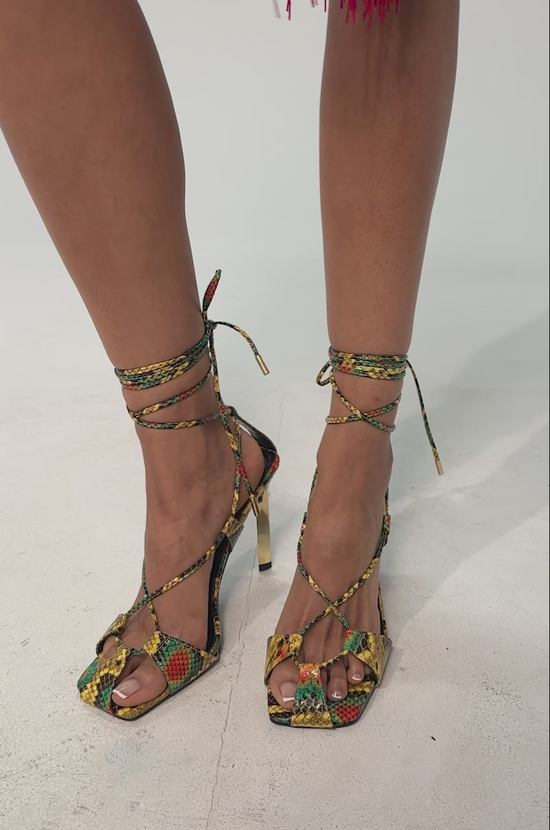 A video of a model wearing the AZALEA WANG NOTYET MULTI SNAKE STILETTO SANDAL which features a yellow, green, and red snakeskin print heel, gold accents, open toe, and lace-up strappy ankle straps.