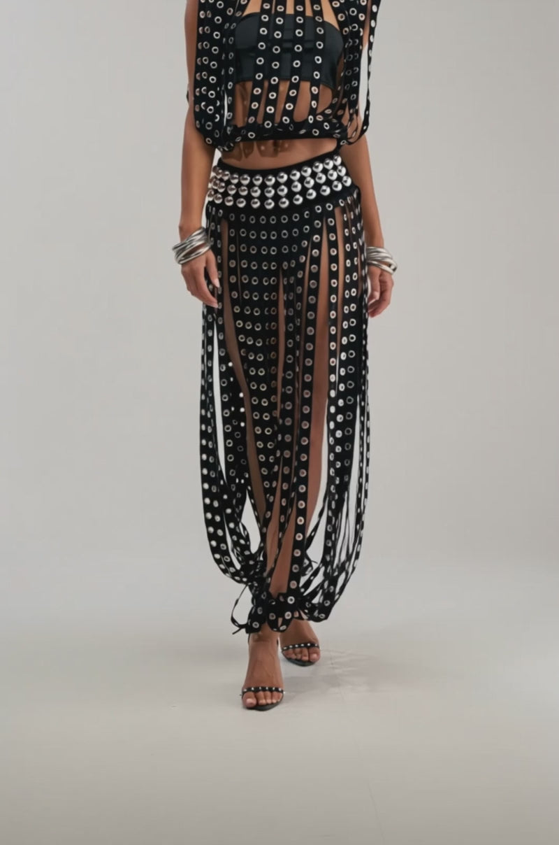 A video of a woman wearing the TOUCH IT GROMMET TOP, a black sheer concert piece with circular cutouts and grommet details, paired with high-waisted bottoms, hoop earrings, silver bangles, and strappy heels.