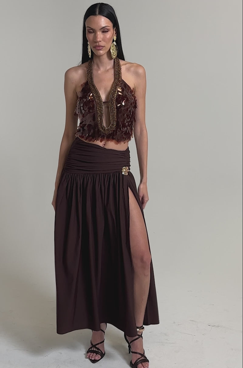 A video of a model wearing the ISLAND GIRL SEQUIN BLOUSE with textured bronze sequins and a matching high-slit skirt, a woman accessorizes with gold earrings and strappy sandals.