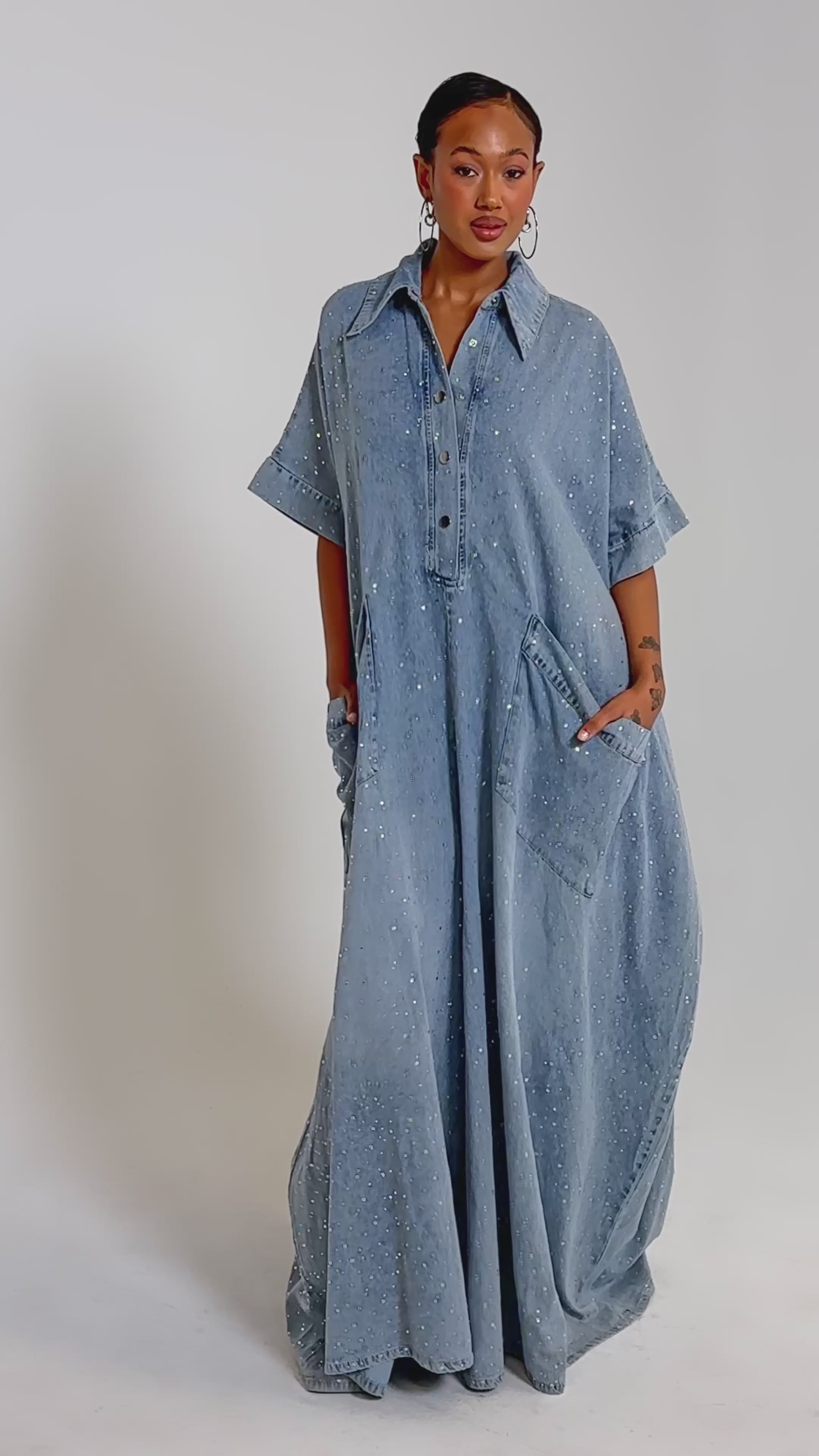 A video of a woman wearing the GINA DENIM RHINESTONE JUMPSUIT with her hands in her pockets against a plain background.