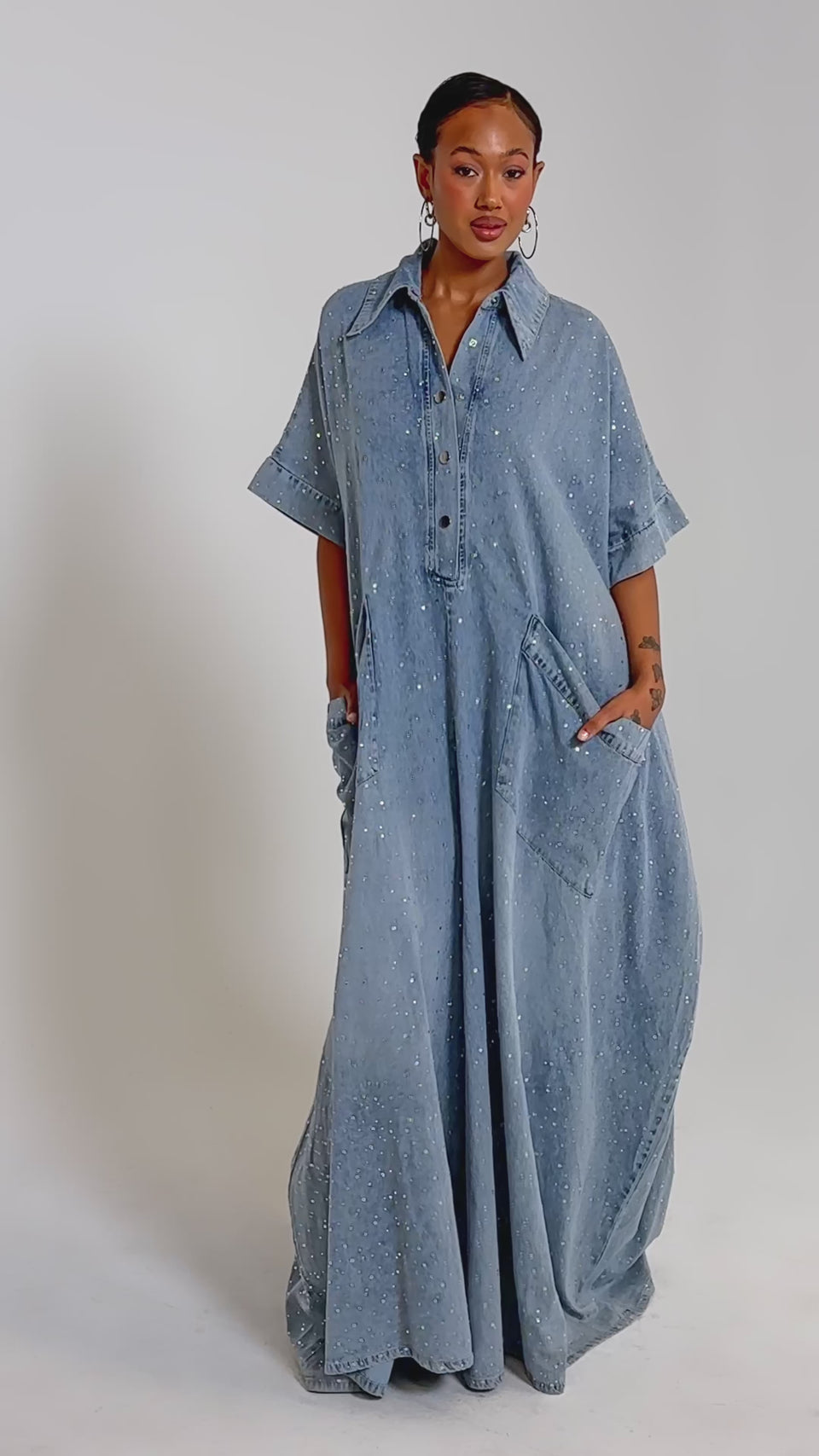 A video of a woman wearing the GINA DENIM RHINESTONE JUMPSUIT with her hands in her pockets against a plain background.
