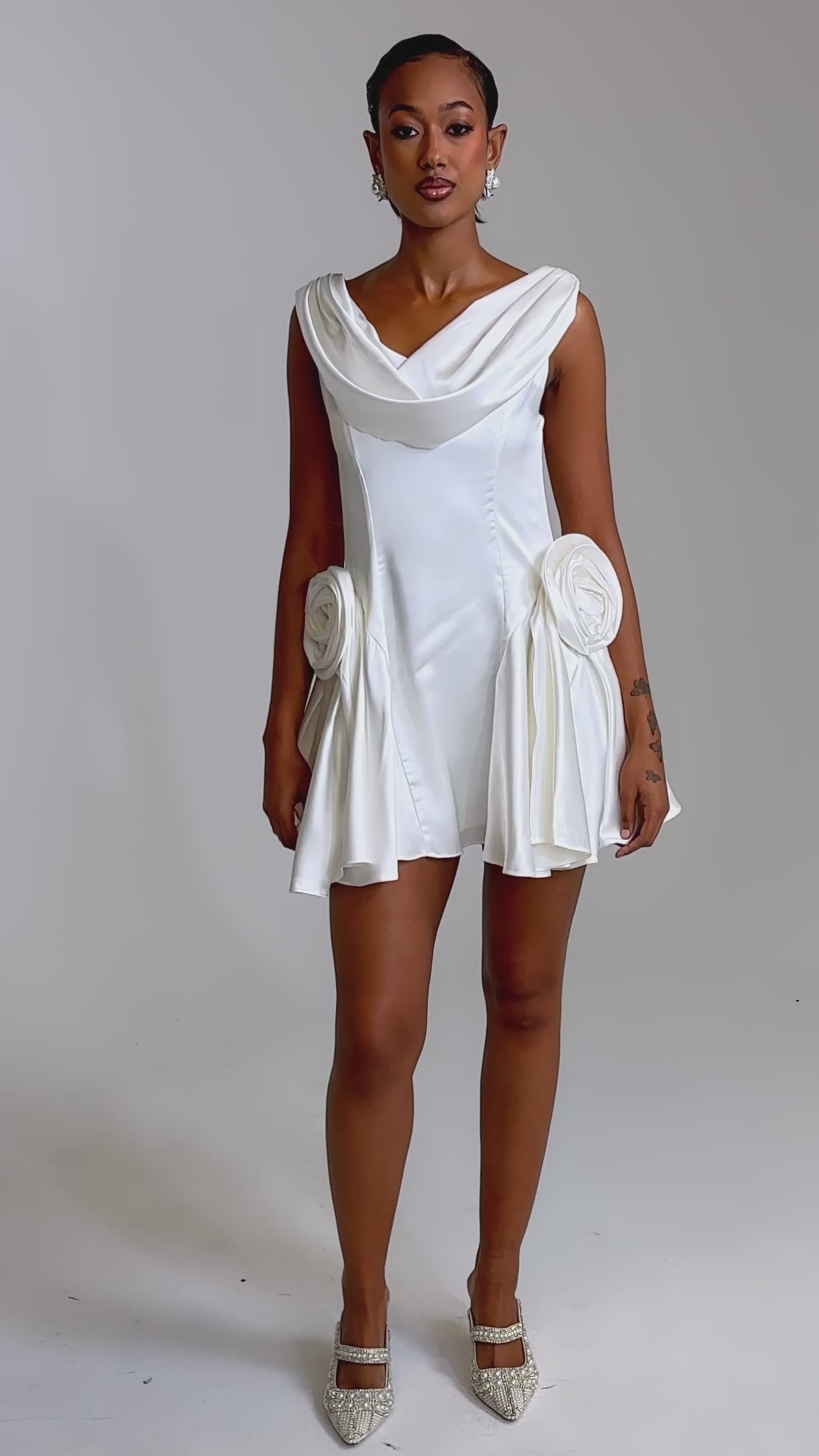 A video of a woman wearing the SWEET ESCAPE MINI DRESS, a short ivory bridal mini with a cowl neckline and large floral accents on each hip. 