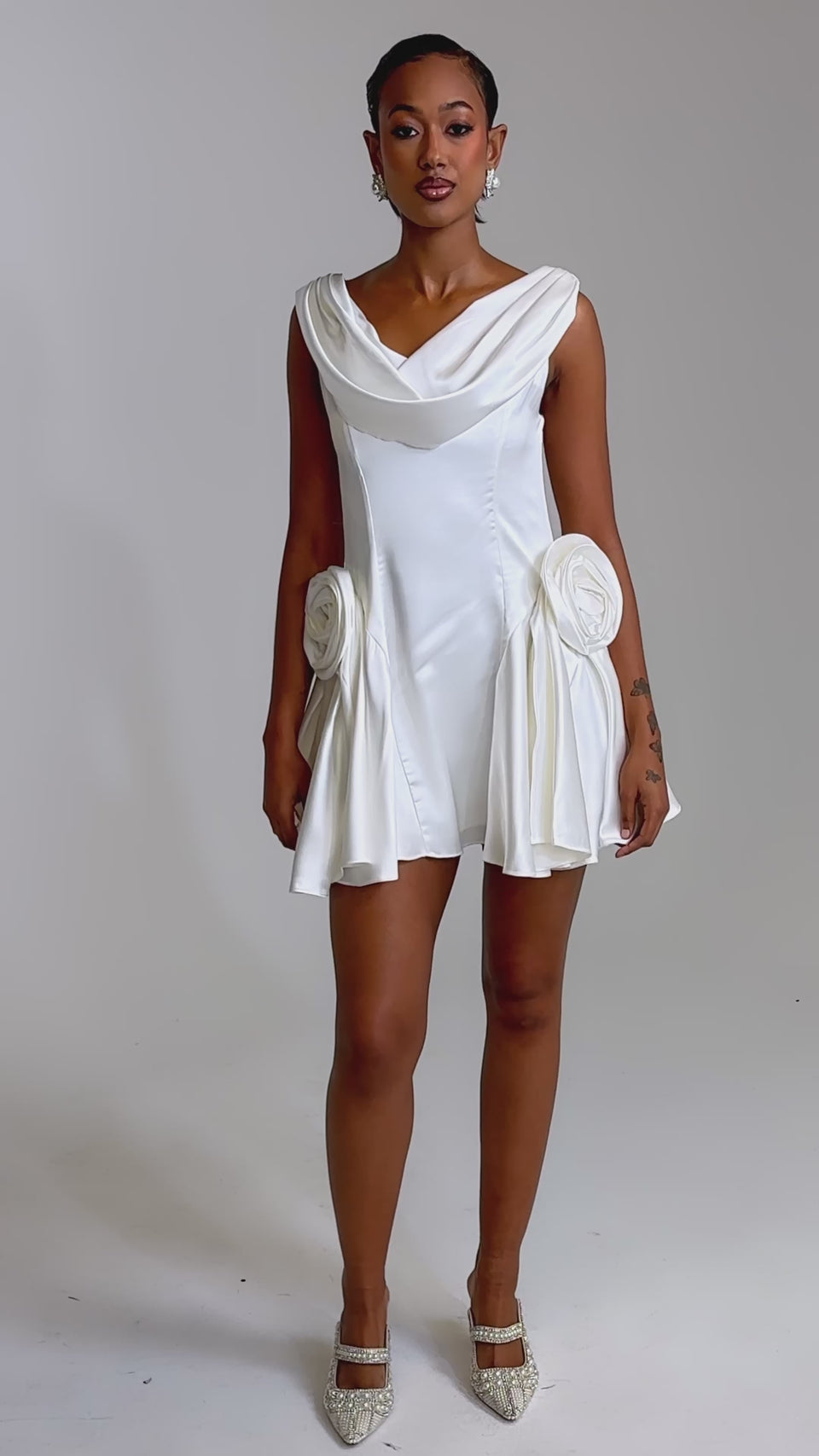 A video of a woman wearing the SWEET ESCAPE MINI DRESS, a short ivory bridal mini with a cowl neckline and large floral accents on each hip. 