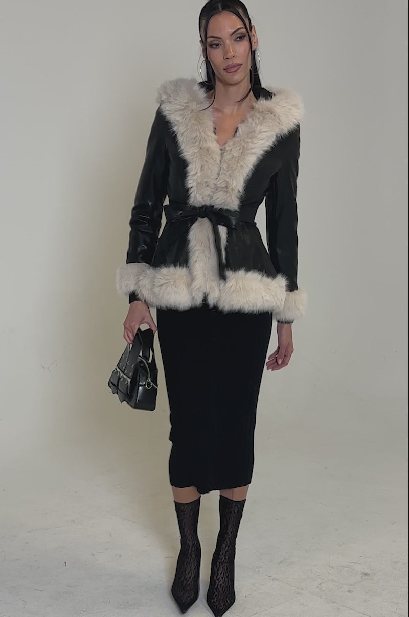 A video of a woman posing in the HOODED KENZY FUR TRIM MOTO jacket with white fur trim, a black ribbed skirt, black lace boots, and a small black handbag against a plain white background.