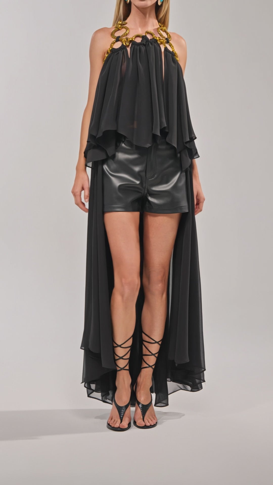 Video of woman modeling the DANCE THE NIGHT AWAY BLOUSE—a black organza top with a high-low silhouette and gold chain neckline—paired with black leather shorts and strappy heels for chic evening style against a plain background.