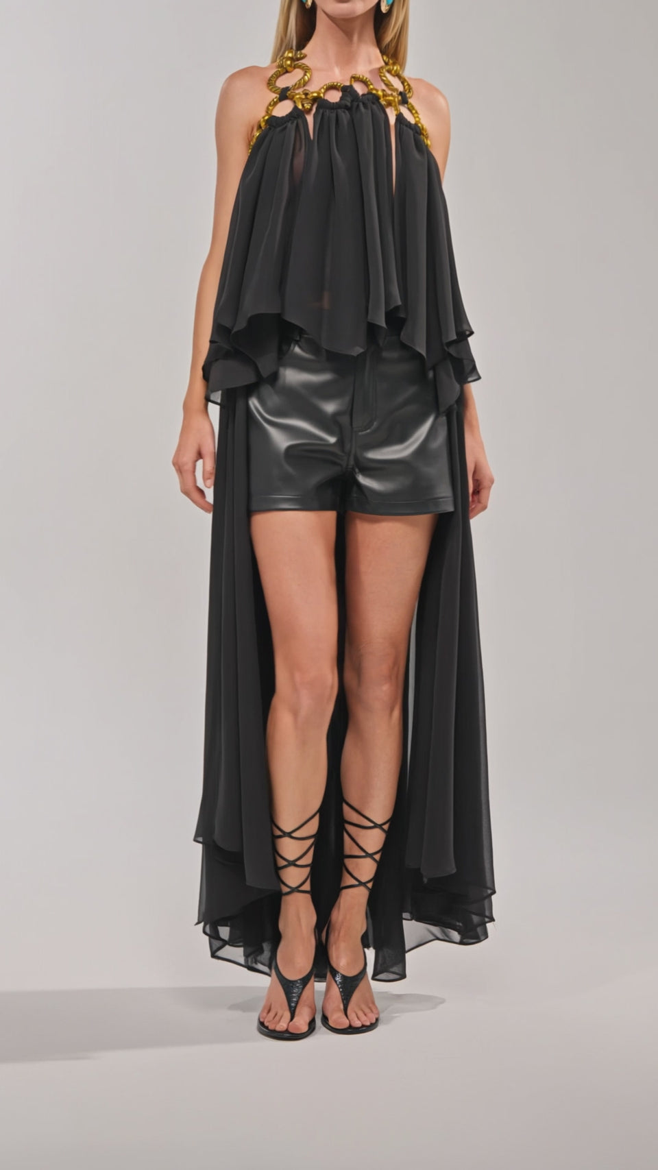 Video of woman modeling the DANCE THE NIGHT AWAY BLOUSE—a black organza top with a high-low silhouette and gold chain neckline—paired with black leather shorts and strappy heels for chic evening style against a plain background.