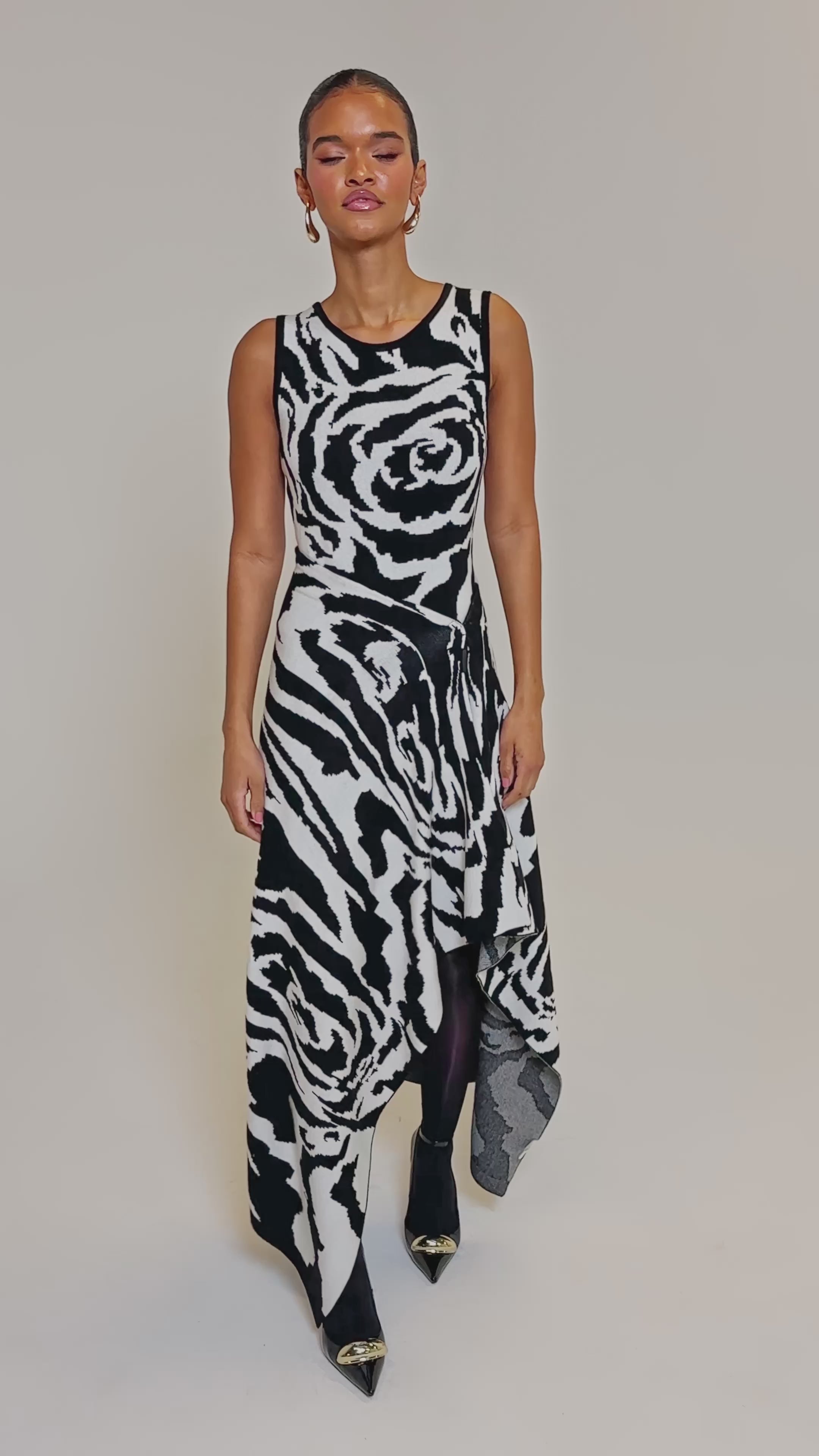 Video of woman modeling the NATOMA FLORAL MAXI SKIRT featuring bold black and white rose and zebra-like prints with an asymmetrical hem, styled with black sheer tights and pointed black heels with gold tips against a plain background.