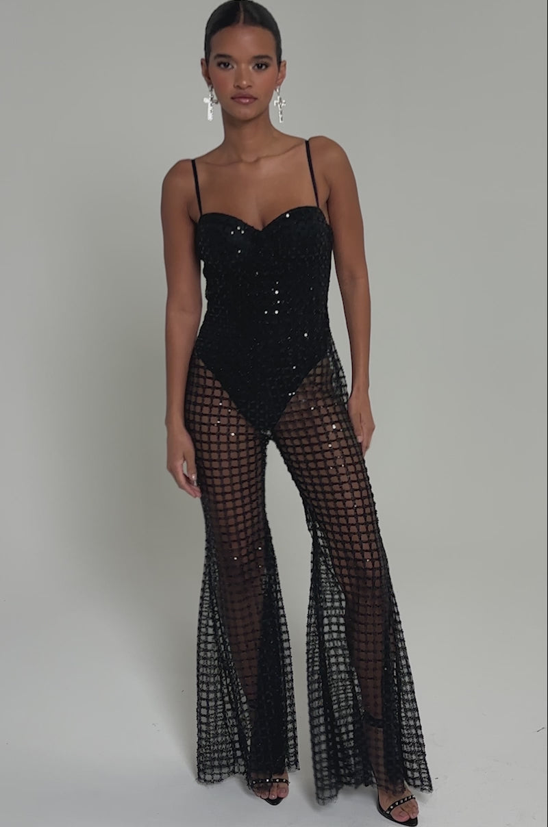 Video of woman modeling the HARDEST TO LOVE MESH JUMPSUIT—black, strapless, with sheer flared pants, a checkered pattern, and sparkling sequin accents—set against a plain light background.