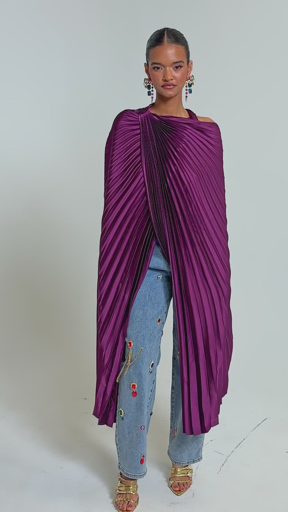 Video of woman modeling the FARAH DRAPED PLEATED CAPE TOP in deep purple with wide sleeves, styled with embellished light blue jeans and gold open-toe heels.