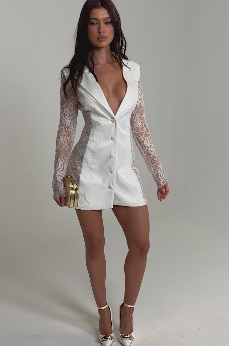 Video of woman modeling the READ ALL ABOUT IT POPLIN JUMPSUIT, featuring sheer lace long sleeves and lace side panels, and holding a gold clutch. She has long brown hair and stands against a plain light background.