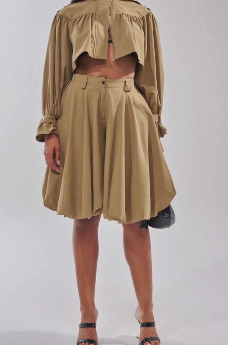 A video of a woman in the CANT STOP WONT STOP BUTTON DOWN, a loose-fitting, tan pleated blouse paired with high-waisted wide shorts. She completes the look with sunglasses, black sandals, and a black handbag.