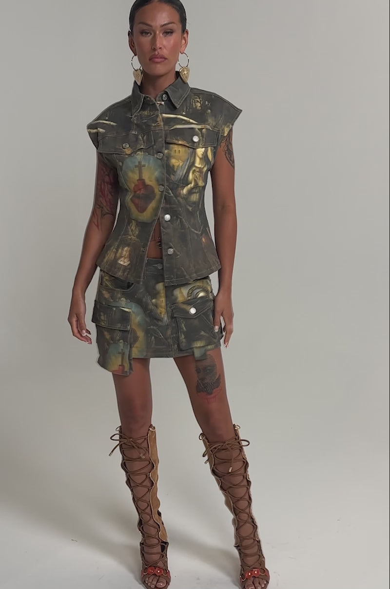 Video of woman modeling the AMEN TO THAT BUTTON DOWN denim top with a baroque print and matching skirt, styled with slicked-back hair, bold leaf earrings, and visible arm tattoos for a striking streetwear festival look.