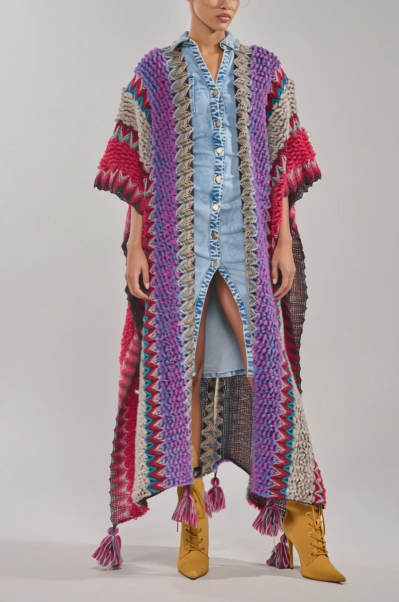 A video of a person wearing the JUST WHAT I AM KIMONO—a colorful, striped boho kimono with tassels at the hem—layered over a blue denim button-down dress, paired with yellow boots and clear eyeglasses.