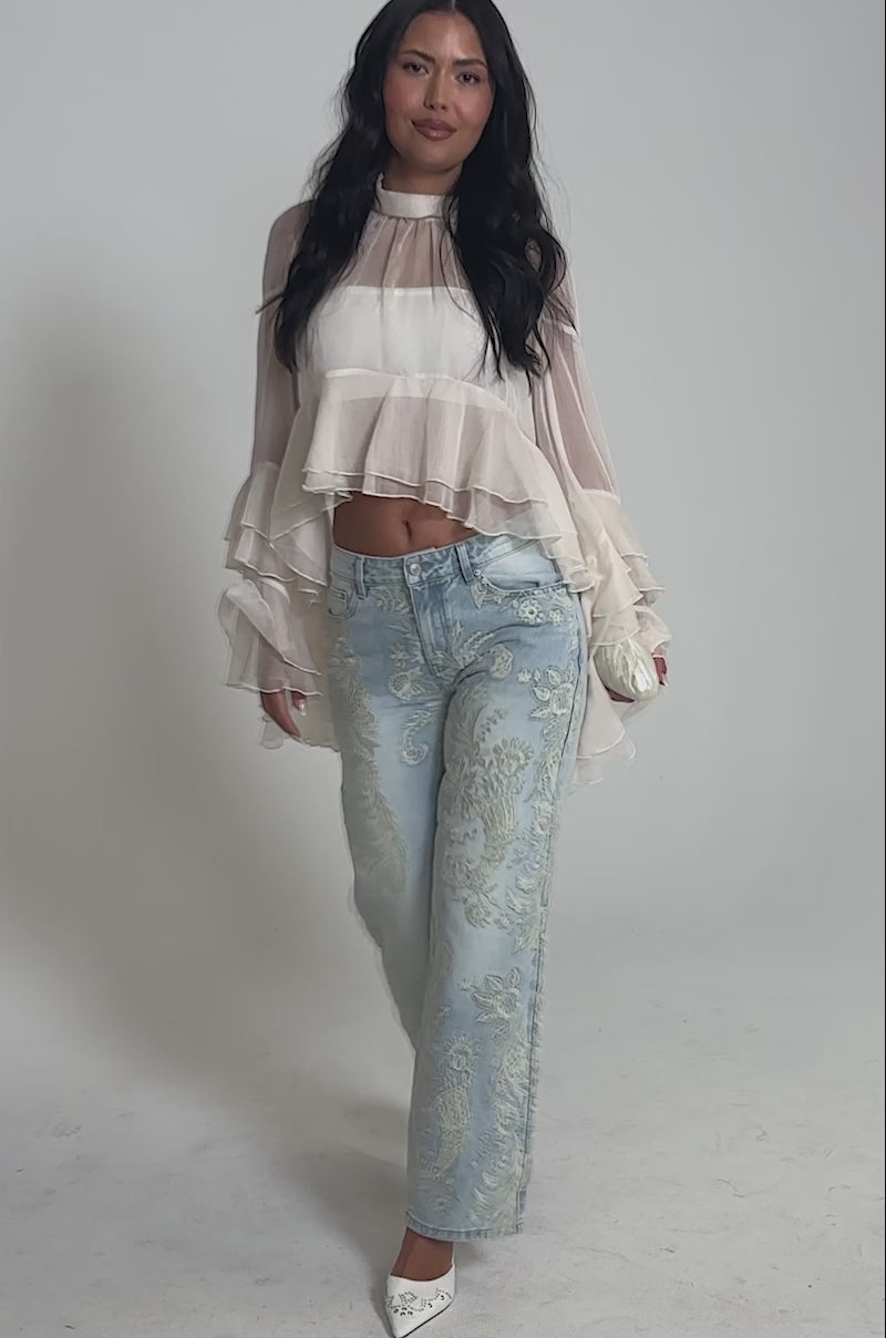 Video of woman modeling the MARGOT CHIFFON BLOUSE, a sheer, tiered ruffle top with bell sleeves, paired with light blue patterned pants. She stands against a plain background and smiles softly.