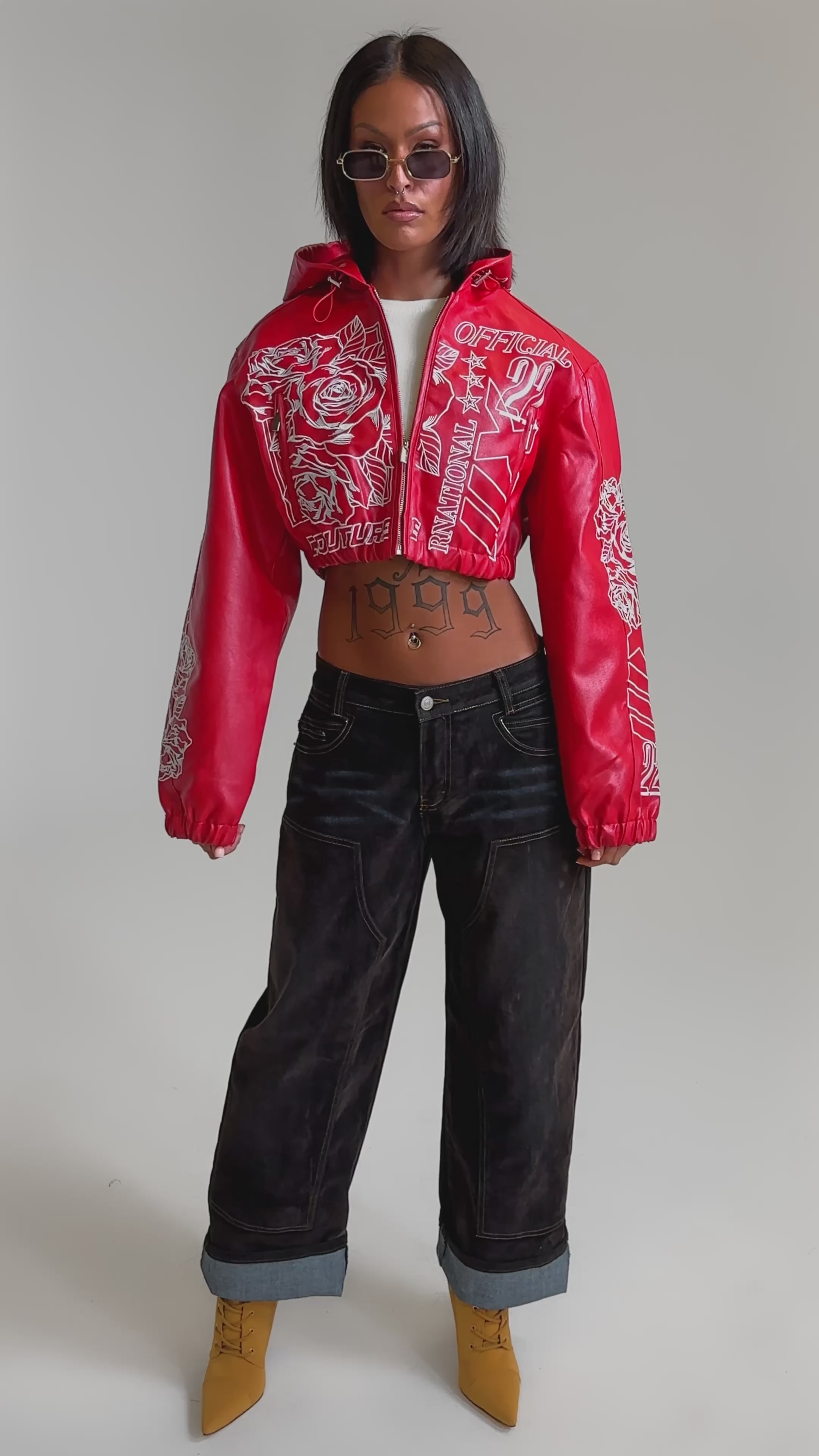 A video of a woman in the PAINT THE TOWN RED EMBROIDERED BOMBER with white designs, baggy dark jeans, and yellow heeled boots. 