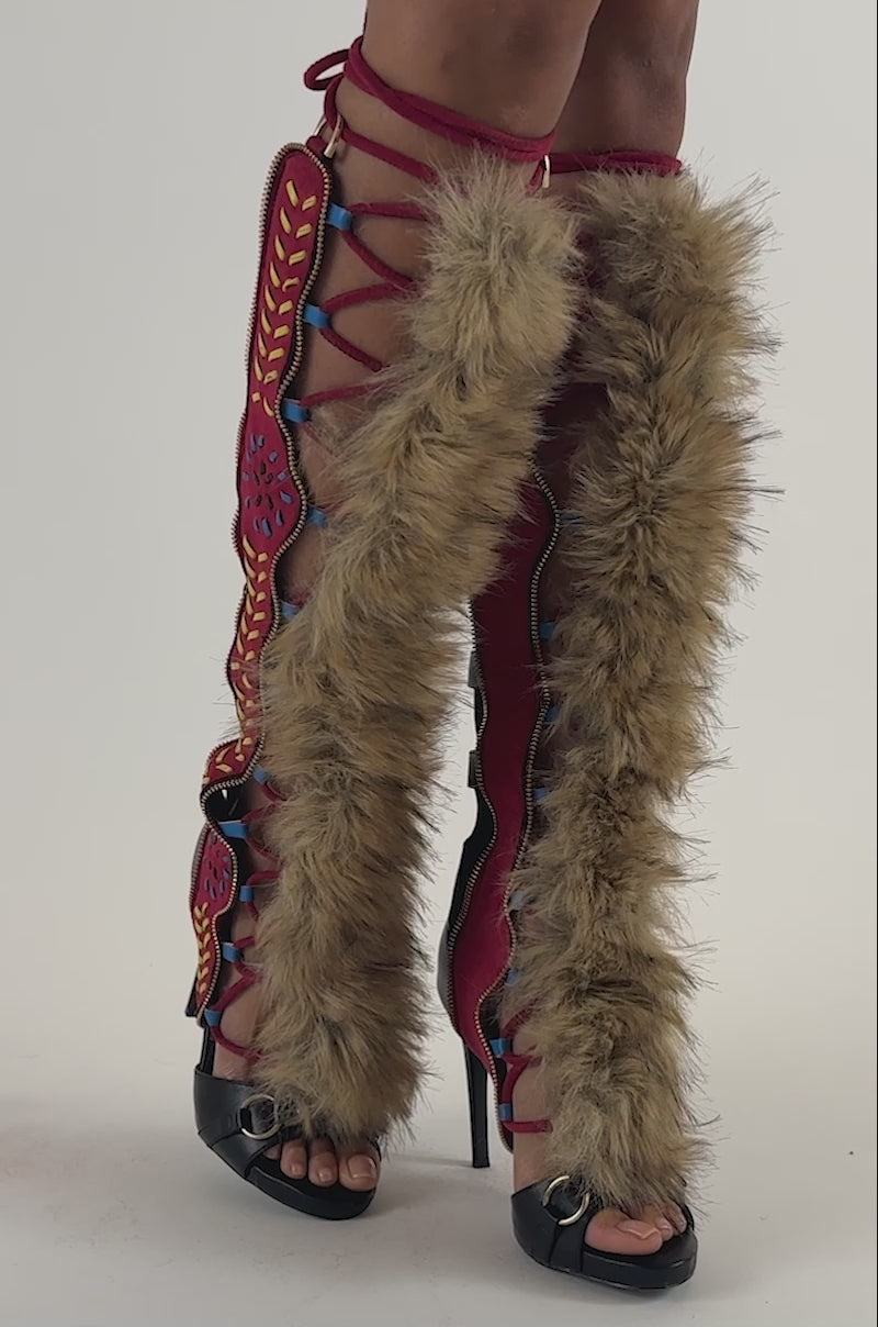 Video of a woman modeling the AZALEA WANG SHAINE BLACK MULTI GLADIATOR STITCH AND FUR SANDALS—distinctive red knee-high lace-up footwear featuring genuine fur trim, blue accents, and embroidered maroon decorative patterns.