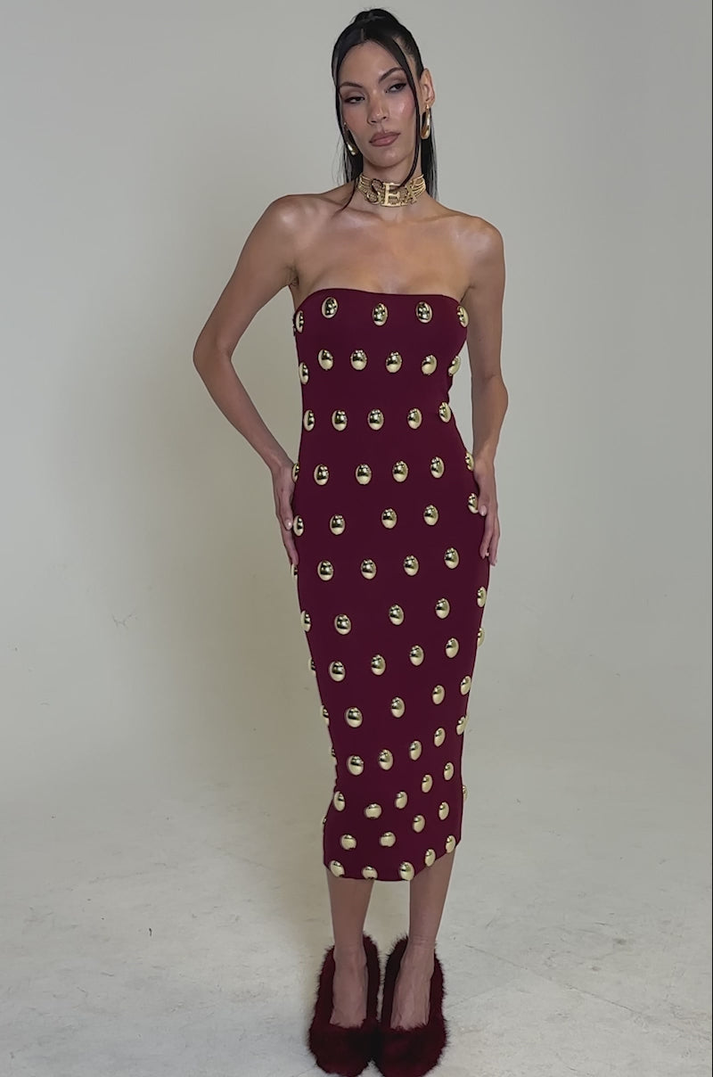 A video of a woman modeling the TIME AFTER TIME MIDI DRESS WITH METAL DETAILING IN BURGUNDY, styled with a chunky gold choker and maroon fuzzy heels, posing against a plain white background.