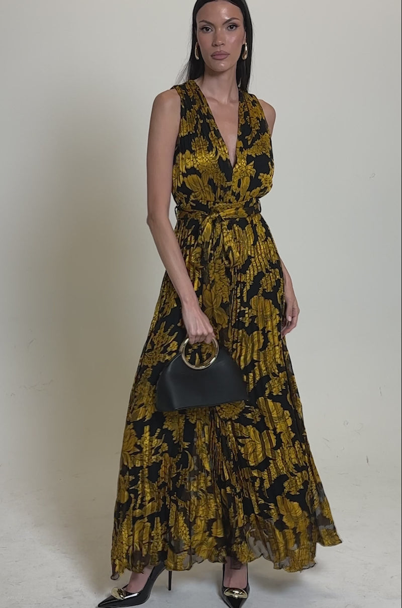 A video of a woman wearing the AKIRA Label REGINA PRINTED WIDE LEG JUMPSUIT, featuring a sleeveless design, V-neckline, yellow floral pattern, tied waist, and wide legs.