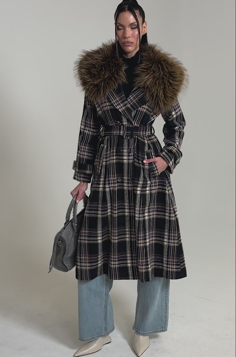 A video of a woman in the SWEPT UP FUR TRIM TRENCH, featuring a large faux fur collar, paired with wide-leg jeans, white pointed-toe boots, and a gray handbag against a plain background.