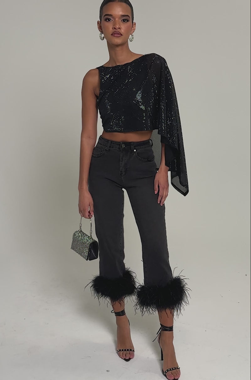 Video of woman modeling the TRIPPY LIKE ME DENIM PANT WITH FEATHER, paired with a black one-shoulder sparkly top, strappy heeled sandals, glittery handbag, and statement earrings. Her hair is styled back for a polished look.