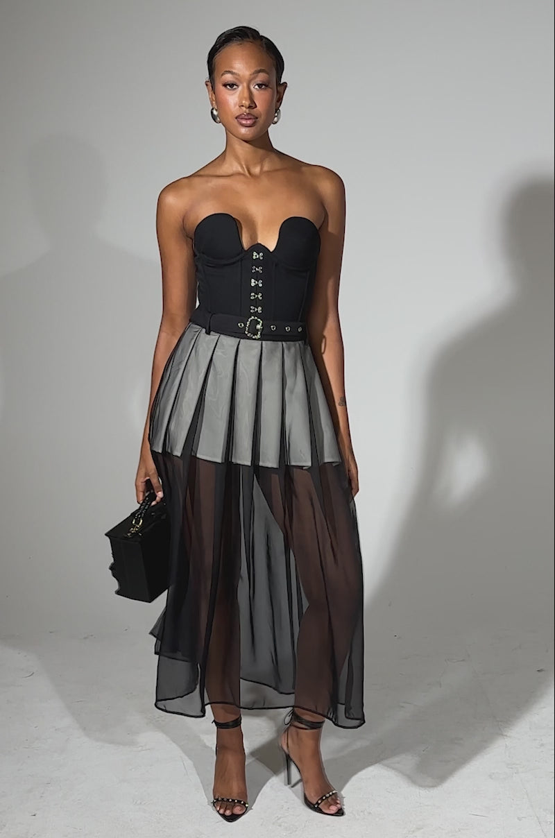 A video of a woman modeling the PARTY GIRL MIDI SKIRT, a sheer mesh, high-waisted midi style. She pairs it with a black strapless corset top, strappy heels, a black handbag, and wears her hair sleeked back against a plain light background.