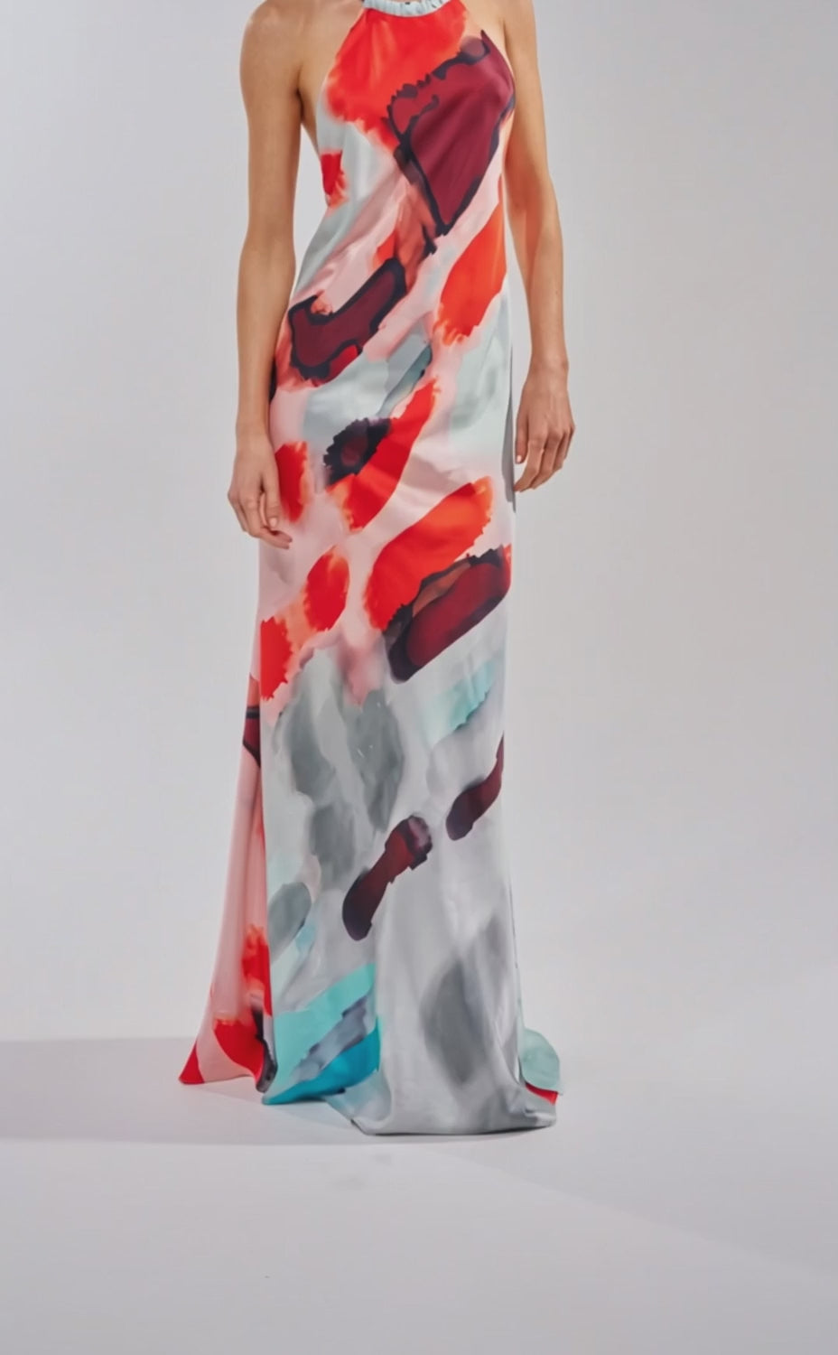 A video of a woman wearing the CHIC IS ME SATIN MAXI DRESS IN MULTI, a flowing halter dress with bold red, black, turquoise, and gray abstract patterns.