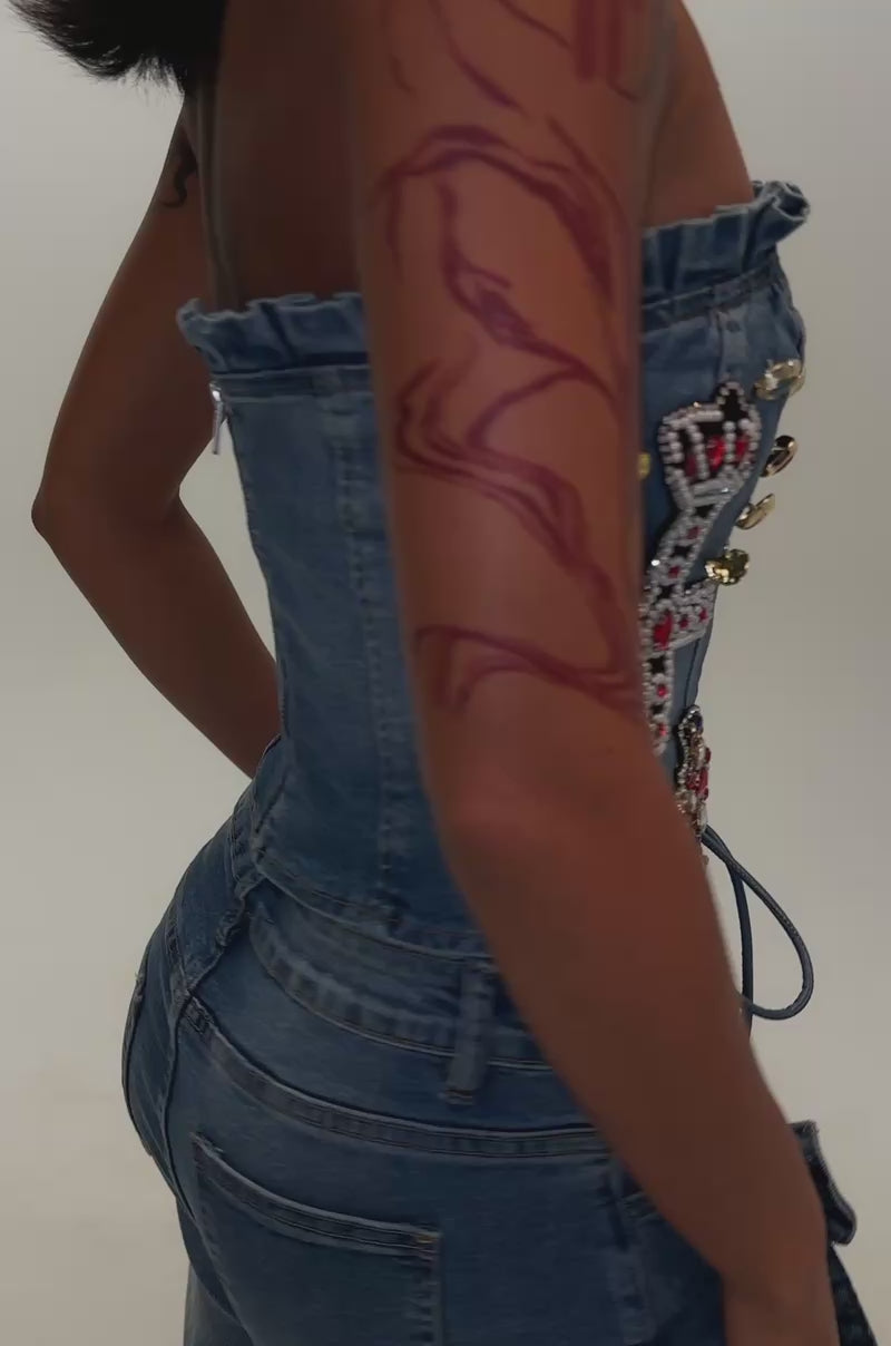 Video of a woman modeling the FEELING BLESSED EMBELLISHED DENIM CORSET IN MEDIUM BLUE, paired with matching jeans, both adorned with colorful jeweled patches, bows, and lace-up details. 