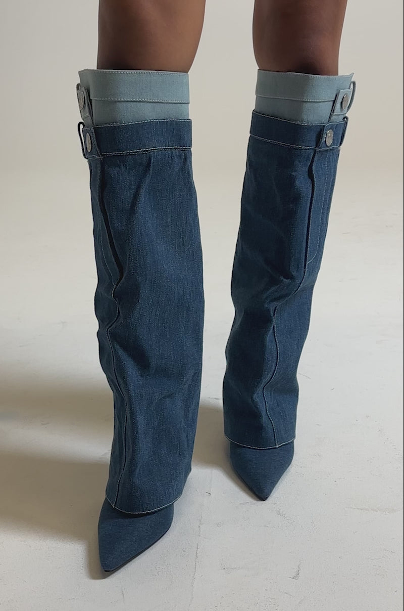 A video of a person wearing the AZALEA WANG DOUBLE FLY DENIM CHUNKY BOOT—unique knee-high denim boots with pant leg details, cuffs, and buttons—standing against a plain gray background.